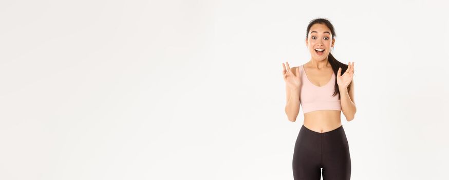 Sport, wellbeing and active lifestyle concept. Happy enthusiastic fitness girl, asian sportswoman clasp hands from fantastic news, gasping amazed, standing over white background