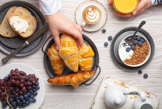 Beautiful morning breakfast gathering with fresh croissants, granola with yoghurt and berries, toasts, coffee cappuccino and fruit on plate. Tasty and healthy breakfast, white wooden table, top view .