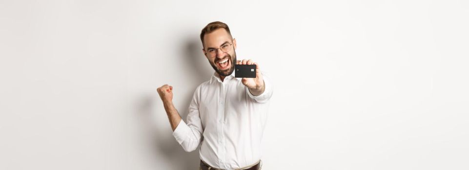 Image of satisfied businessman showing credit card, making fist pump in rejoice, standing over white background