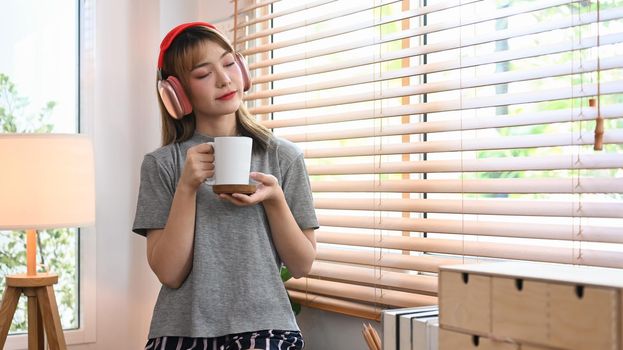 Calm young woman holding cup of coffee and listening yo music on wireless headphone, enjoy stress free peaceful mood