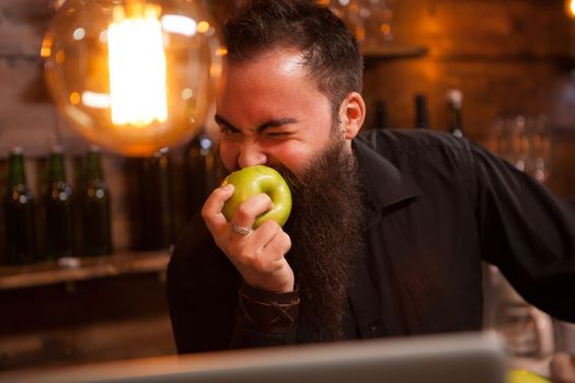 Bearderd bartender eating a green apple in vintage pub