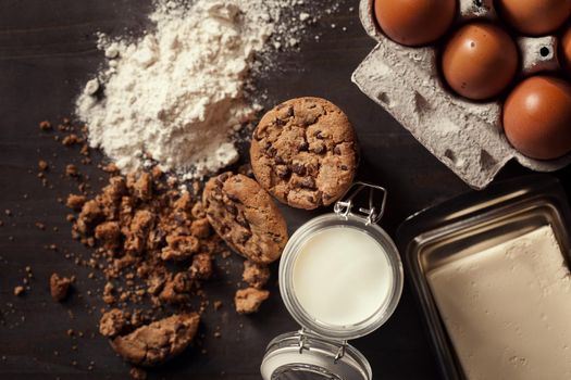 Flat lay of Homemade chocolate chip cookies with bottle of milk, white flour, fresh eggs,butter and crumbs on rustic wooden table.