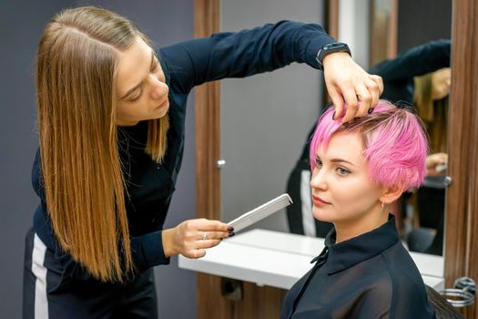 Female hairdresser styling short pink hair of the young white woman with hands and comb in a hair salon.