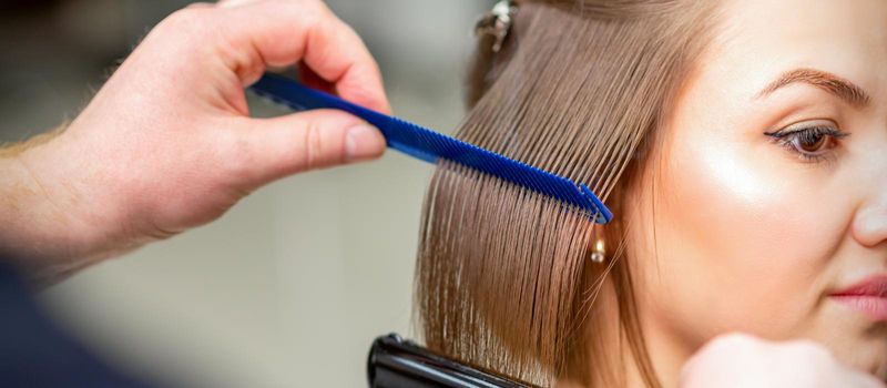 Hairdresser straightens female brown hair of medium length with a hair with an iron hair straightener and comb in a beauty salon.