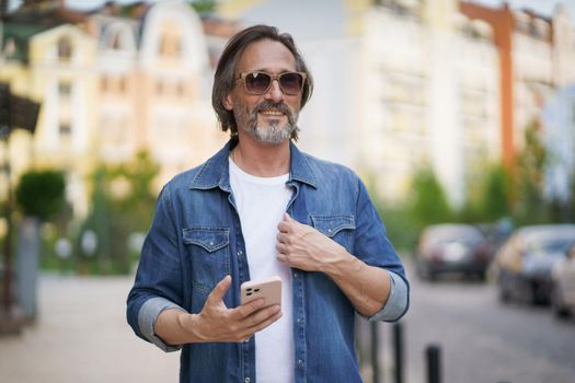 Mature handsome man with smartphone in hand walking on old town streets wearing denim shirt and sunglasses. Handsome middle aged man with grey hair making photos while traveling