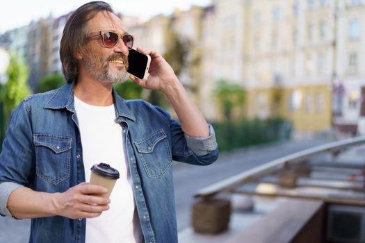 Handsome grey beard middle aged man talking on the phone holding coffee in disposable paper cup standing outdoors in old city background wearing jeans shirt. Freelancer traveling man