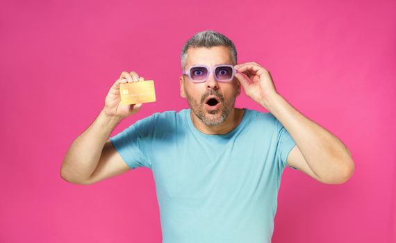 Excited of a new offer handsome man hold debit, credit card in hand lowering his blue glasses wearing blue t-shirt isolated on pink background. Man with bank card in hand. Financial, banking concept