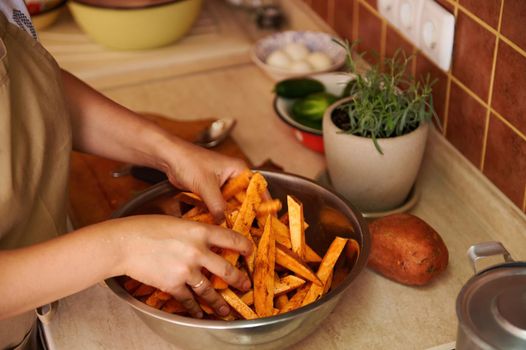 Close-up housewife standing by kitchen table and seasoning sliced wedges of organic batata with fragrant culinary herbs