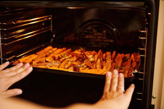 Close-up chef's hands putting a baking sheet with raw organic sweet potatoes wedges into an oven