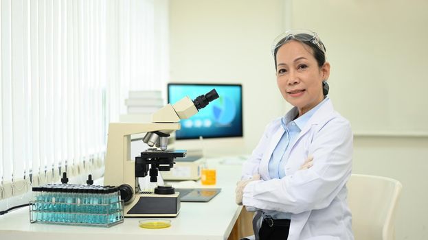 Middle age woman wearing scientist uniform sitting with arms crossed at laboratory