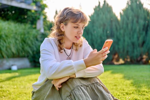 Cute teenage girl sitting with smartphone on grass in backyard