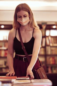 Covid, face mask and woman with books in a library, bookshop or educational building for knowledge, reading or research. Young female student browsing and choosing novel on table in a bookstore