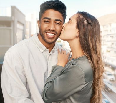Happy couple, love and kiss on cheek from girlfriend bonding with boyfriend standing on a city building balcony on a holiday. Portrait of indian man and beautiful woman in interracial relationship