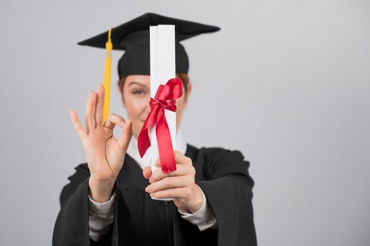 Happy woman in graduation gown holding out diploma and showing ok sign.