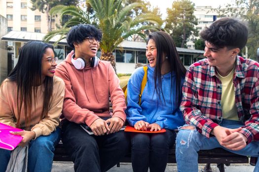 Group of multiracial gen z college student friends talk and laugh sitting on campus bench outside.