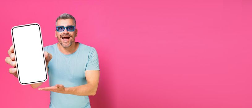 Smartphone in hand of handsome man, guy 30s 40s in casual blue shirt and sunglasses isolated on pink background. Man with phone studio shot. Mobile app advertisement. Copy space
