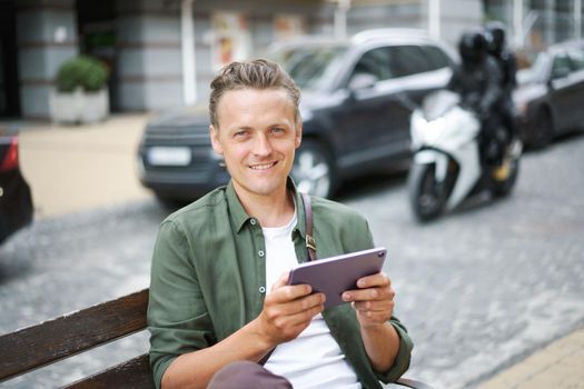 Portrait of young freelancer man work using digital tablet sits on the bench outdoors in front office building wearing casual green shirt and shoulder bag. Young handsome man working on the go