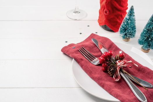 Christmas table setting with white dishware, silverware and red and green decorations on white wooden background