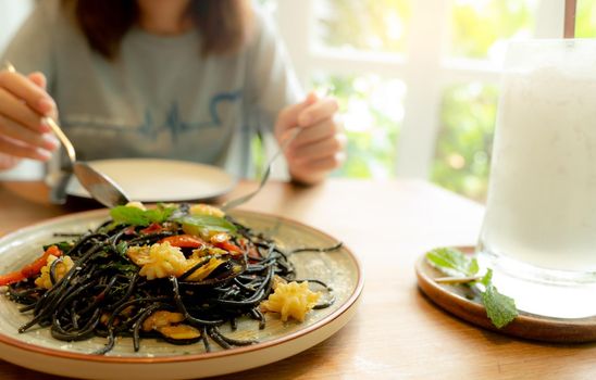 Black cuttlefish ink spaghetti with shrimp on plate. Black pasta with squid ink on a restaurant table and blur woman eating with fork and spoon. Healthy food. Woman eating delicious black spaghetti.