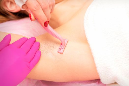 Beautician shaves the armpit of a young woman with a razor before the hair removal procedure.