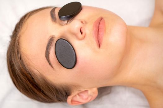 Young woman receiving facial massage with black mineral stones on her eyes in a spa.