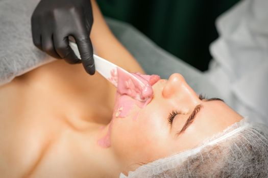 Beautician applying alginate peel-off powder facial mask with the spatula in a spa.