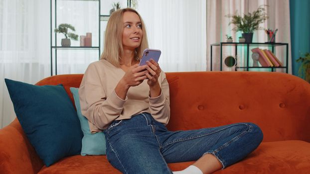Portrait of adult girl sitting on sofa, using smartphone share messages on social media application