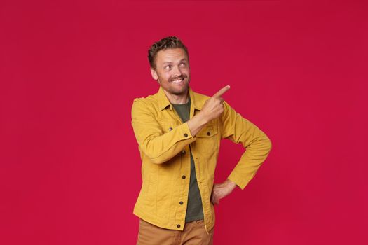 Handsome young man in jeans jacket pointing looking sideways up mock up advertisement isolated on red background. Charming smiling freelancer introduce your business offer