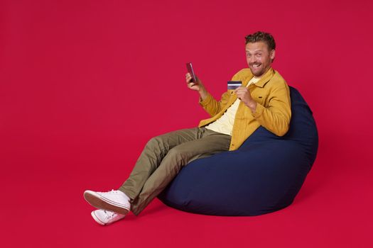 Buying online using debit, credit card handsome young man 30s wearing casual denim clothes sit in bag chair holding digital tablet in hands working isolated on red background. Freelancer man