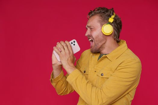 Handsome man 30s singing enjoying his favorite song or track using phone and wireless headphones wearing denim yellow jacket isolated on red background. Joyful man sing while listen music
