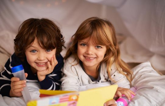 Live, love and learn together. two adorable siblings using torches to read a book inside their blanket fort.
