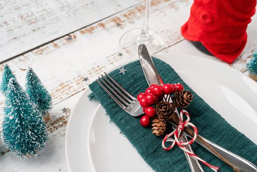 Christmas table setting with white dishware, silverware and red and green decorations on white wooden background. High angle view.