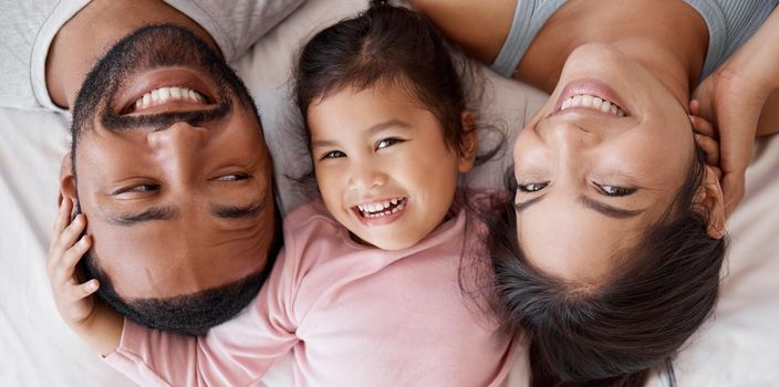 Happy family with child on bed in a face portrait for interracial love, care and happiness together. Girl kid from Mexico with mother and father or parents relaxing at home and bonding from above