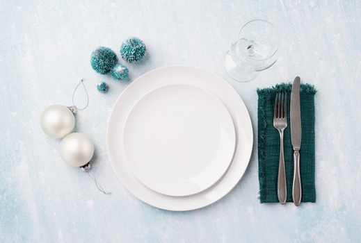 Christmas table setting with white dishware, silverware and green decorations on blue background. Top view.