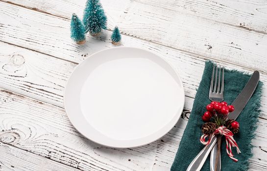 Christmas table setting with white dishware, silverware and red and green decorations on white wooden background