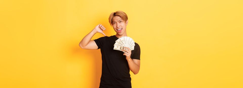 Sassy handsome asian guy pointing finger at cash and looking pleased. Korean man borrowed money, standing yellow background