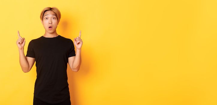 Portrait of astonished asian guy in black t-shirt, open mouth fascinated, pointing fingers up, yellow background