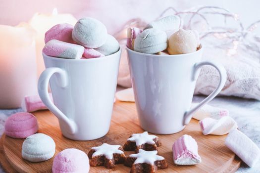 White mugs of hot winter drink with color marshmallow, cookies in the shape of stars, candles and knitted sweater