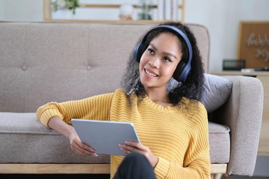 Photo of beautiful dark skin wavy lady homey mood holding digital tablet earflaps listening new audio sitting floor near couch casual clothes yellow at living room indoors