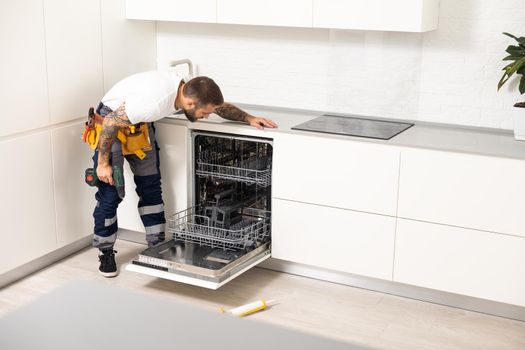 Young modern serviceman in worker suit during the repairing of the dishwasher on the domestic kitchen