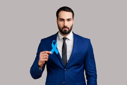 Portrait of serious bearded man holding blue ribbon in hands, awareness prostate cancer.
