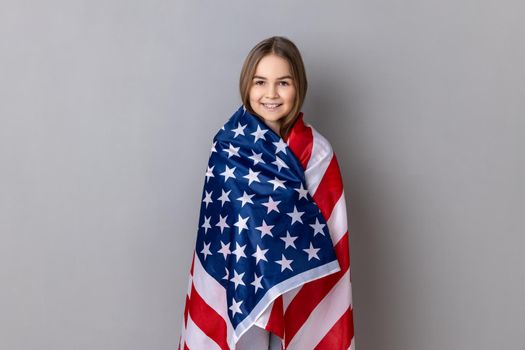 Patriotic little girl standing wrapped in american flag, looking at camera, relocating to America.