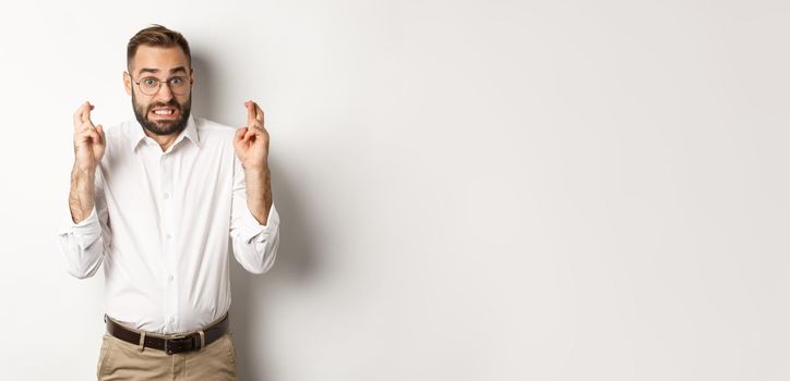 Worried man making a wish, cross fingers and hope for relish, standing over white background