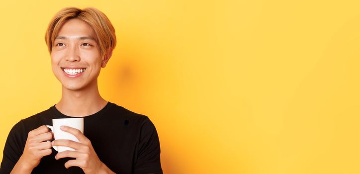 Close-up of happy smiling handsome blond asian guy, looking away with dreamy nostalgic look while drinking coffee, standing over yellow background