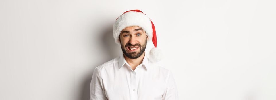 Close-up of awkward guy in santa hat apologizing, feeling uncomfortable, standing over white background. Christmas concept