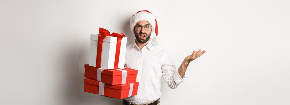 Merry christmas, holidays concept. Man looking confused while holding xmas gifts, shrugging puzzled, standing in santa hat against white background
