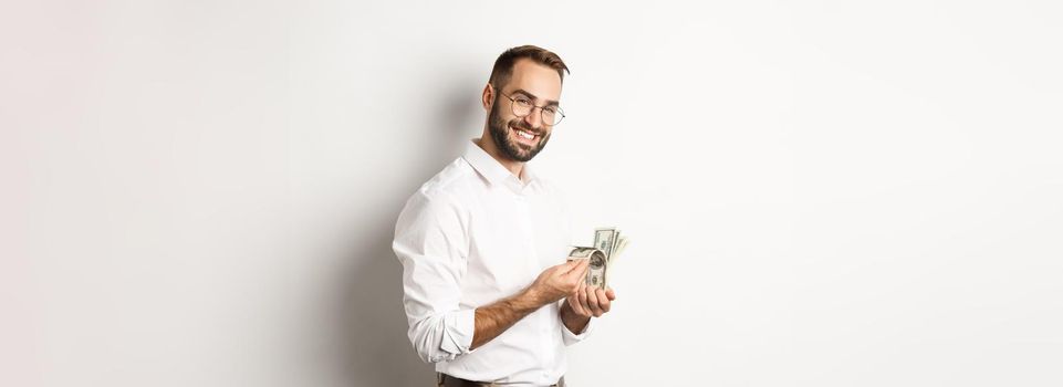 Successful business man counting money and smiling, standing against white background and looking satisfied