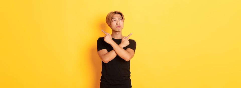 Indecisive handsome asian guy looking confused, pointing fingers sideways, standing yellow background