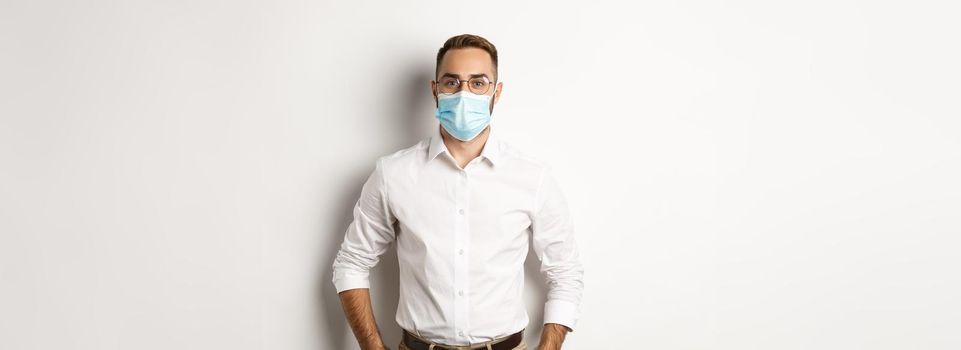 Covid-19, social distancing and quarantine concept. Male employee wearing face mask for work, standing against white background