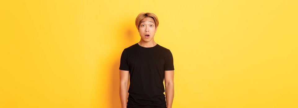 Portrait of startled handsome asian blond guy, drop jaw and gasping impressed, yellow background